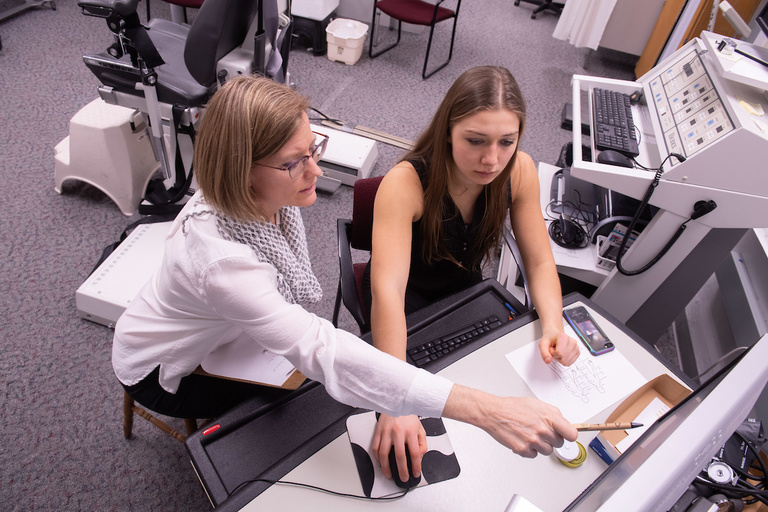 Two women sit at a desk, looking at a computer monitor