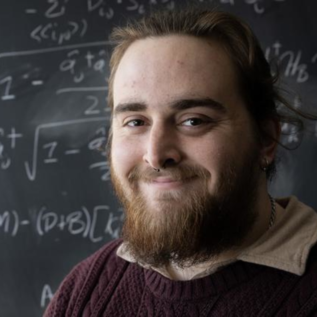 Bearded white man smiles before a black board covered in mathematical formulas