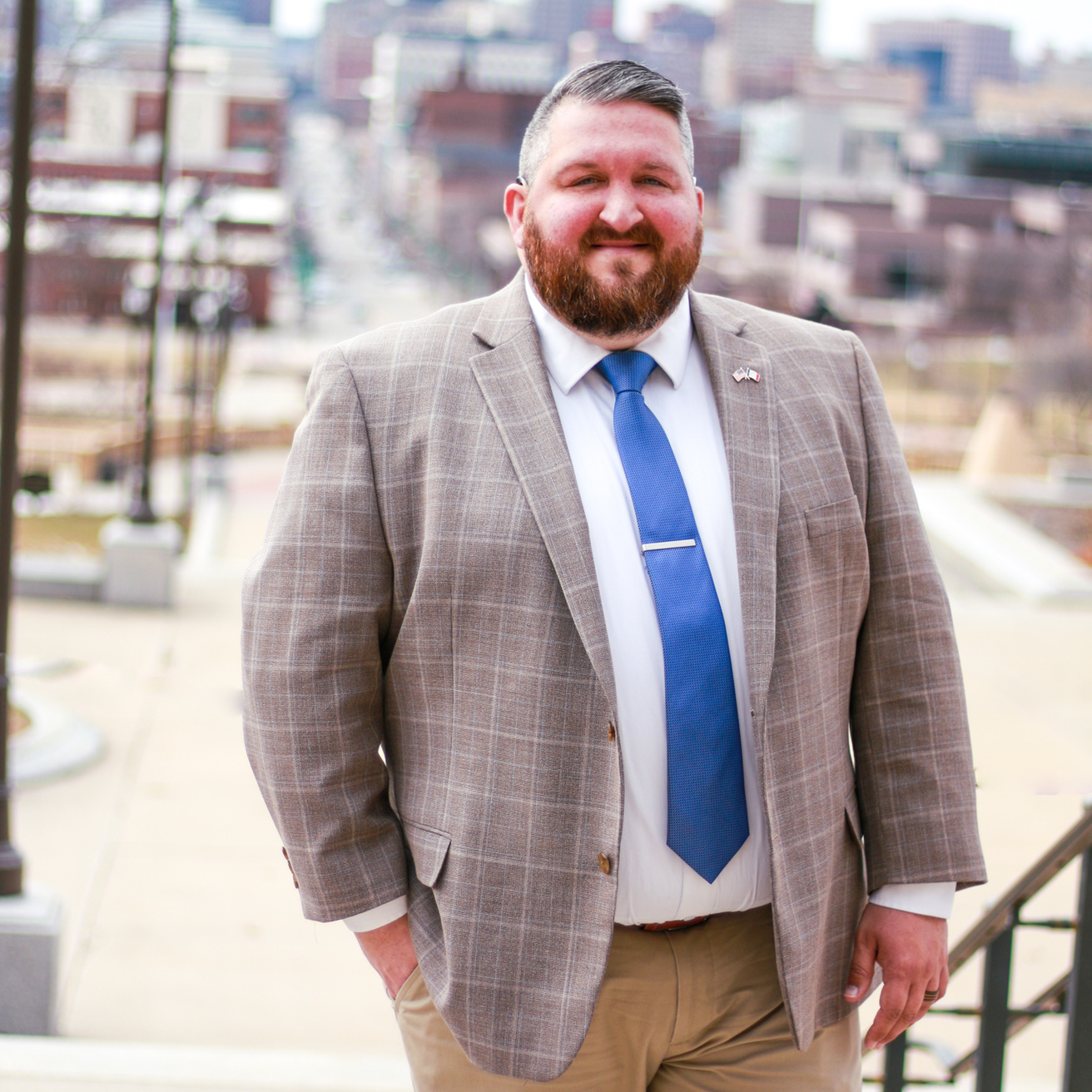 White man in a tweed blazer and blue tie smiles with a downtown cityscape behind him.